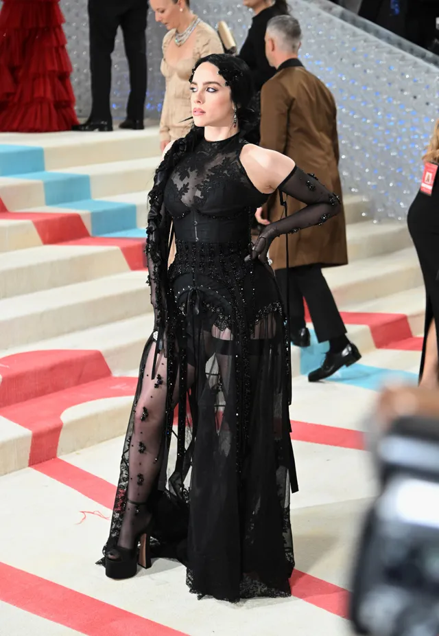 Billie Eilish standing beside colorful event steps in a black gown and heels.