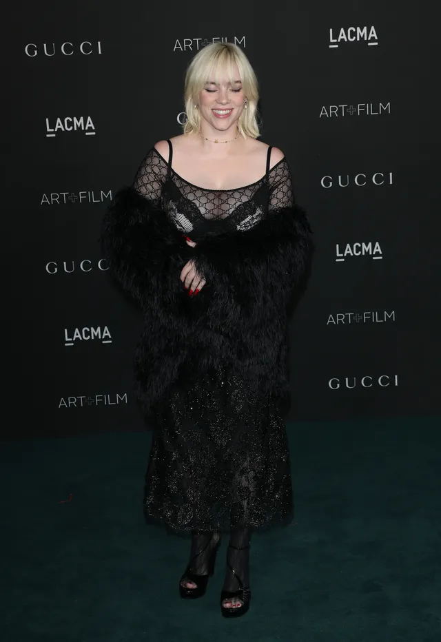 Billie Eilish standing at an event in a lace dress and dark heels.