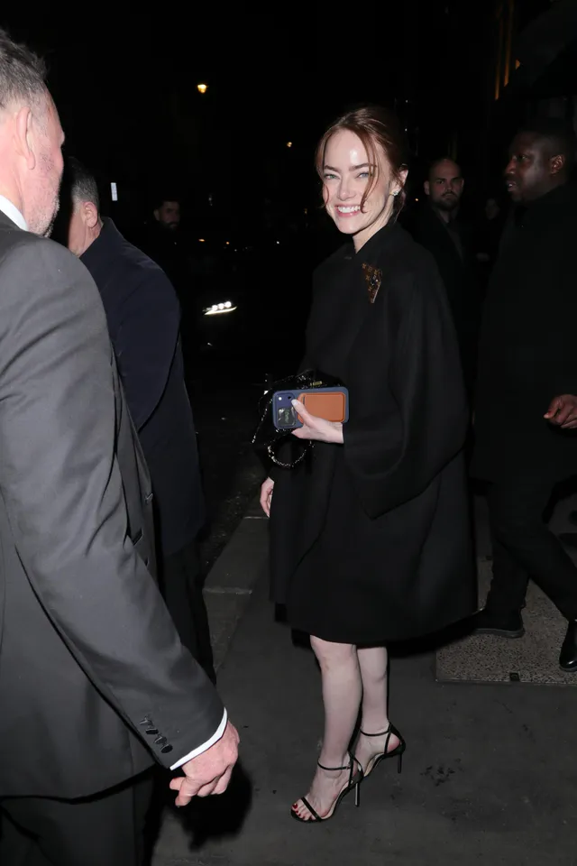 Emma Stone standing among guests in a black cape dress and strappy heels.