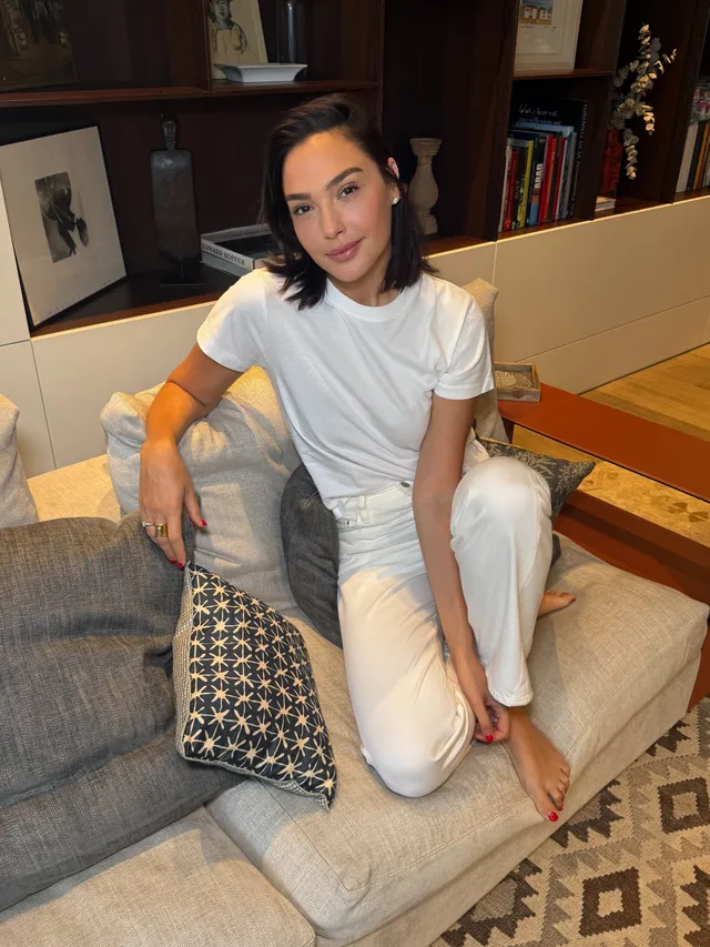 Gal Gadot sitting barefoot on a gray sofa in a white outfit.