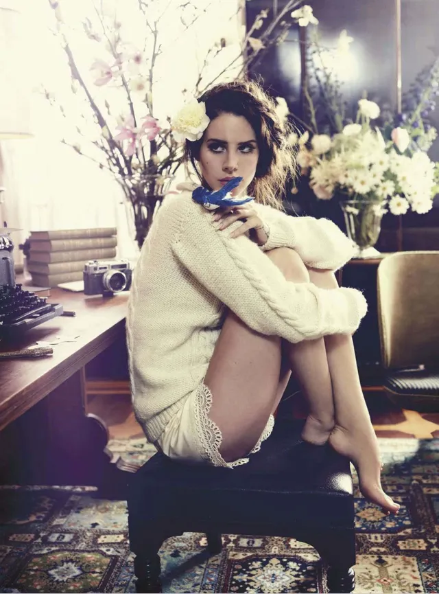 Lana Del Rey sitting barefoot on a swivel chair in a pale knitted outfit.