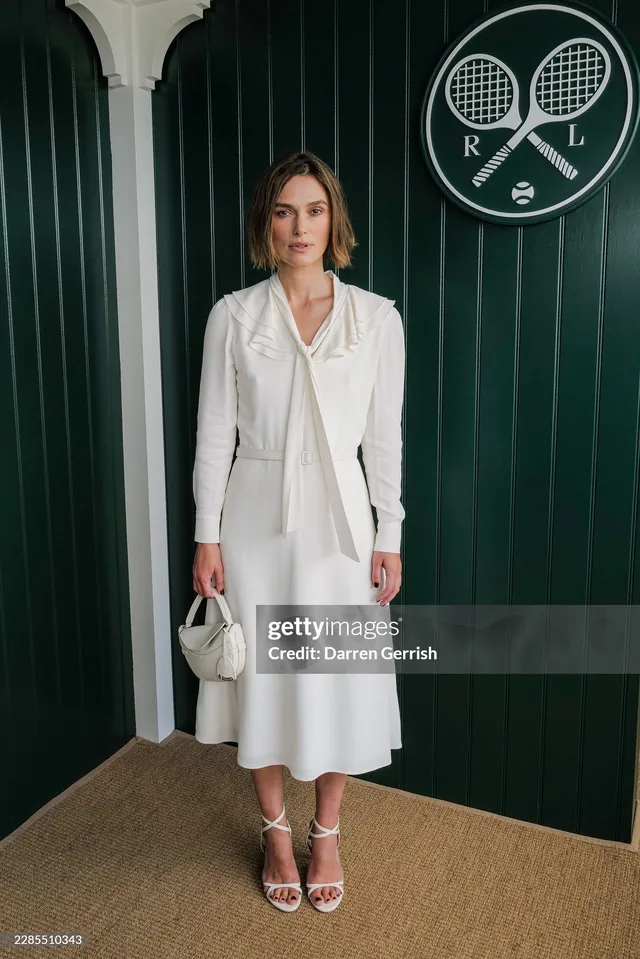 Keira Knightley standing beside a green doorway in a white outfit and heels.