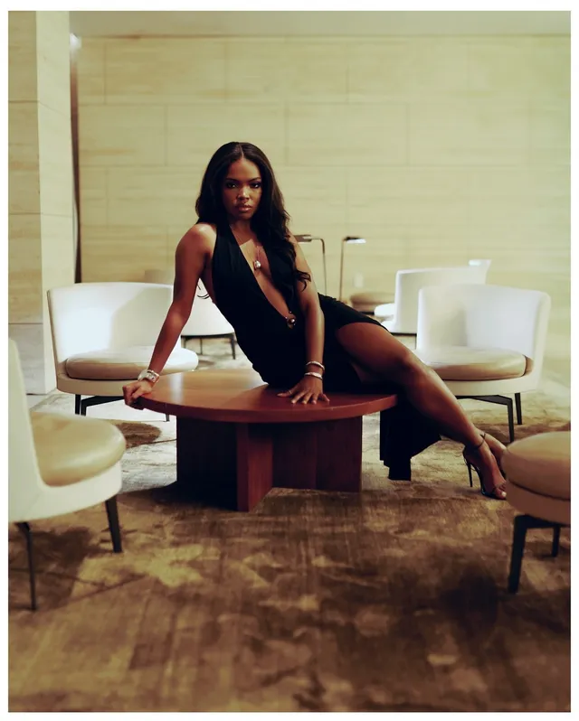 Ryan Destiny sitting on a table in a black dress and heels.