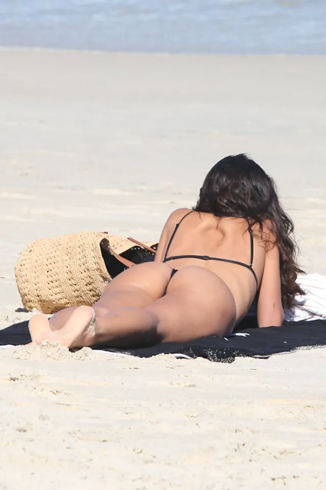 Bruna Marquezine reclining barefoot on a dark beach towel in a bikini.