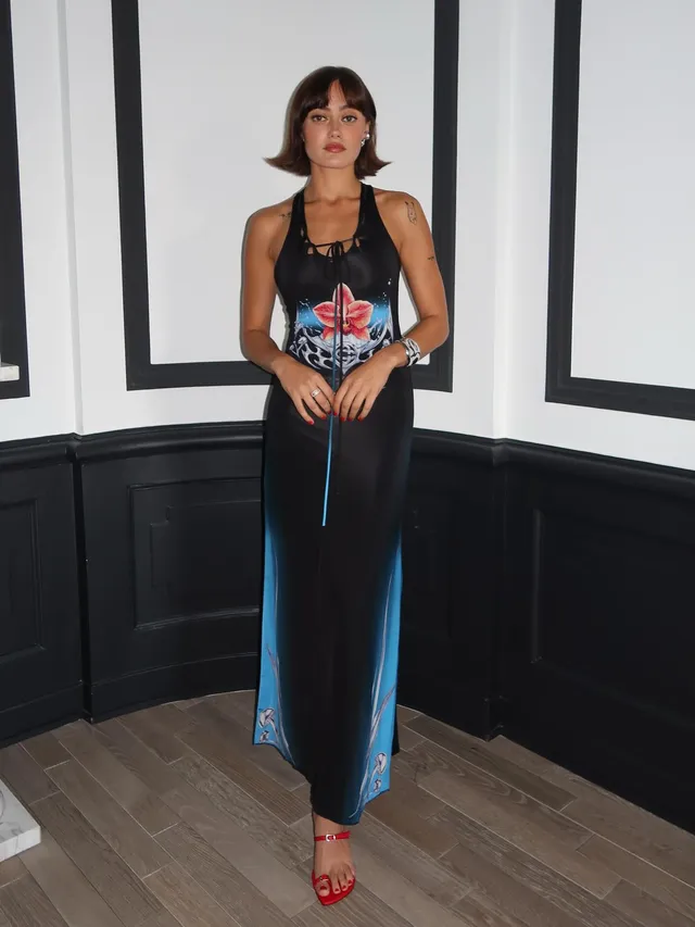 Ella Purnell posing indoors in a dark flowing outfit and red sandals.