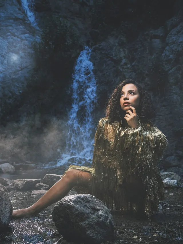Kiana Madeira sitting barefoot on rocks beside a waterfall in a patterned outfit.