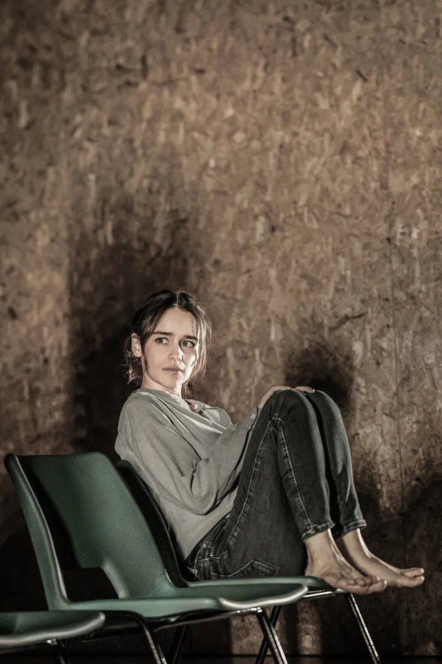 Emilia Clarke reclining barefoot on green chairs for a portrait.