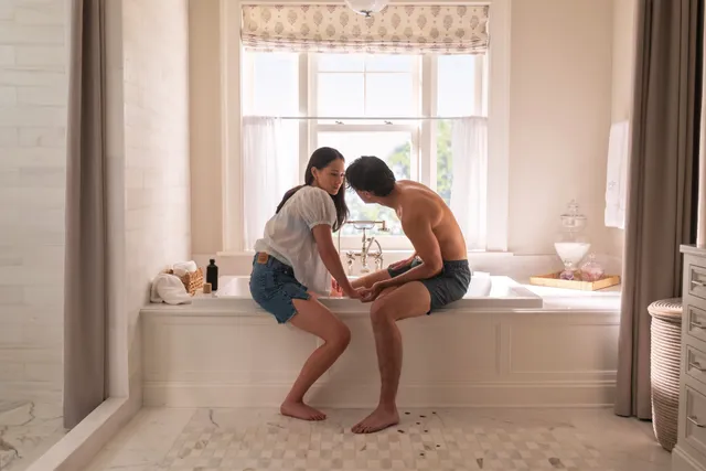 Lola Tung sitting barefoot beside a man on a bathroom ledge.
