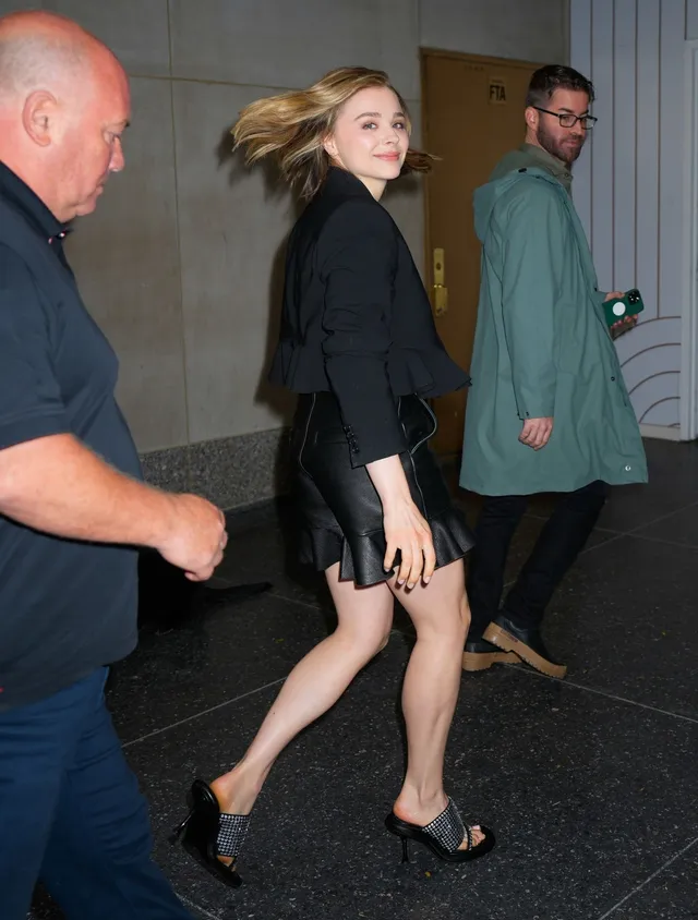Chloë Grace Moretz walking beside other people in a dark outfit and heels.