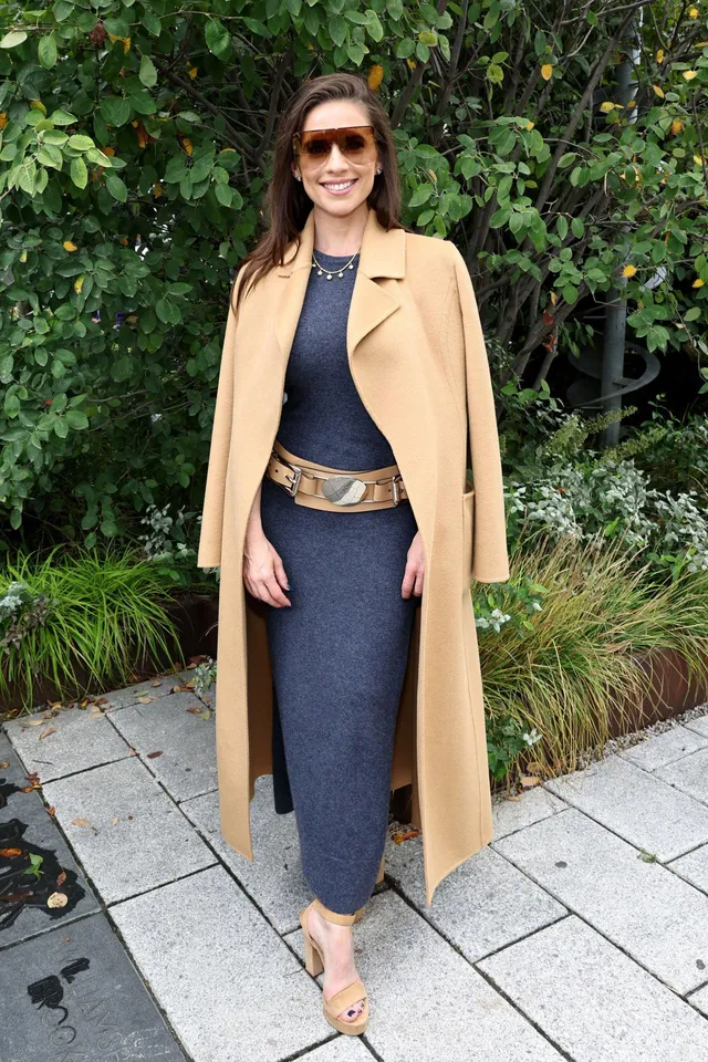 Hayley Atwell standing beside greenery in a camel coat.