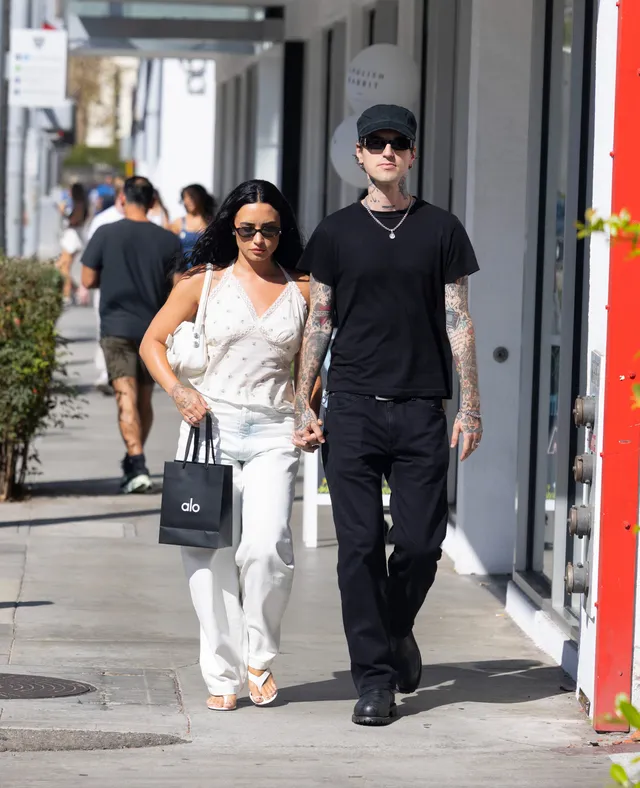 Demi Lovato walking outdoors in white trousers and flat sandals.