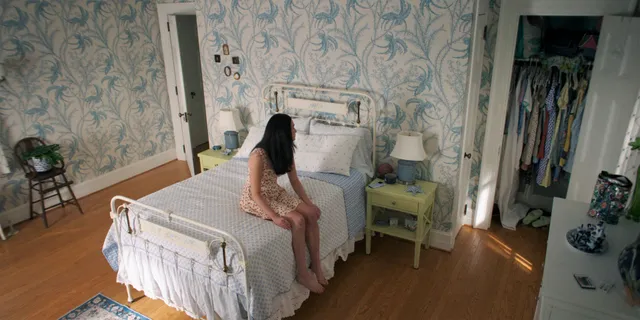 Lola Tung sitting barefoot on the edge of a bed.