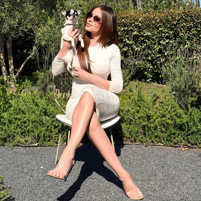 Sofía Vergara sitting barefoot outdoors in a white dress while holding a dog.