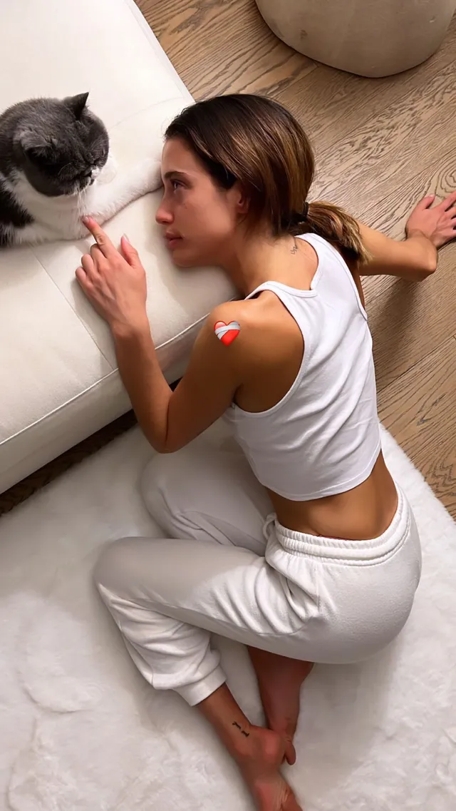 María Pedraza kneeling barefoot beside a sofa and a cat in a white outfit.