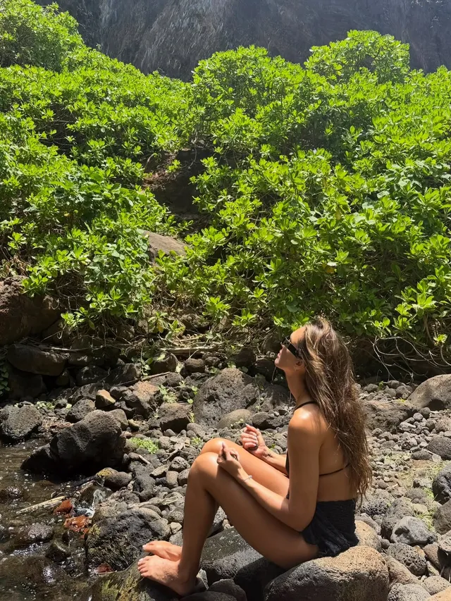 Jessica Alba sitting barefoot on coastal rocks.