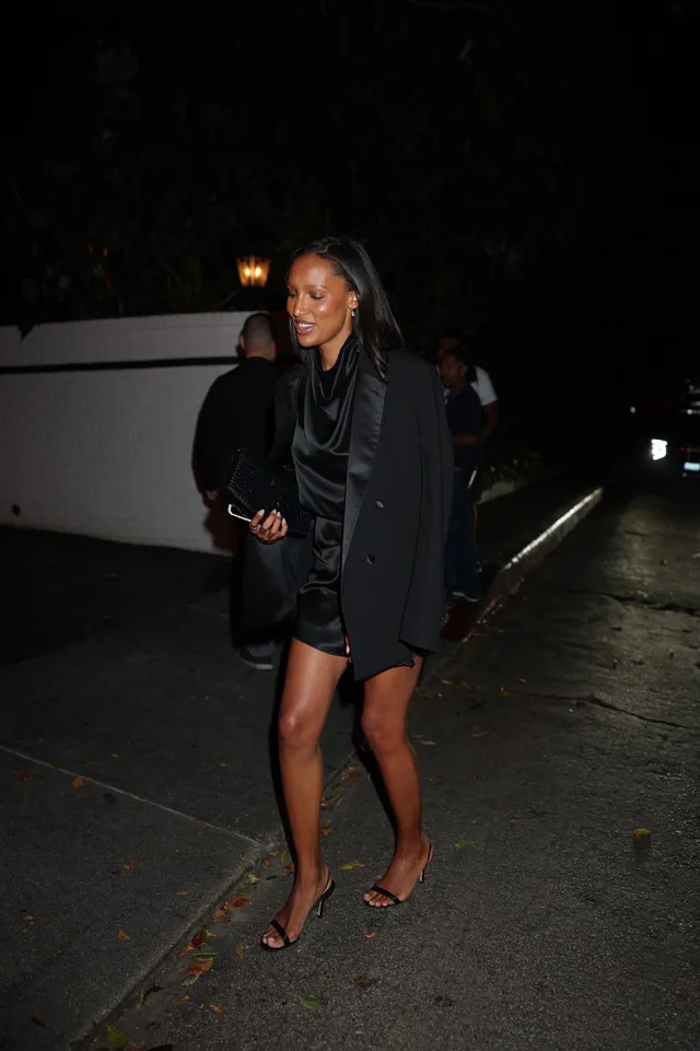 Jasmine Tookes walking outdoors in a black outfit and heeled sandals at night.