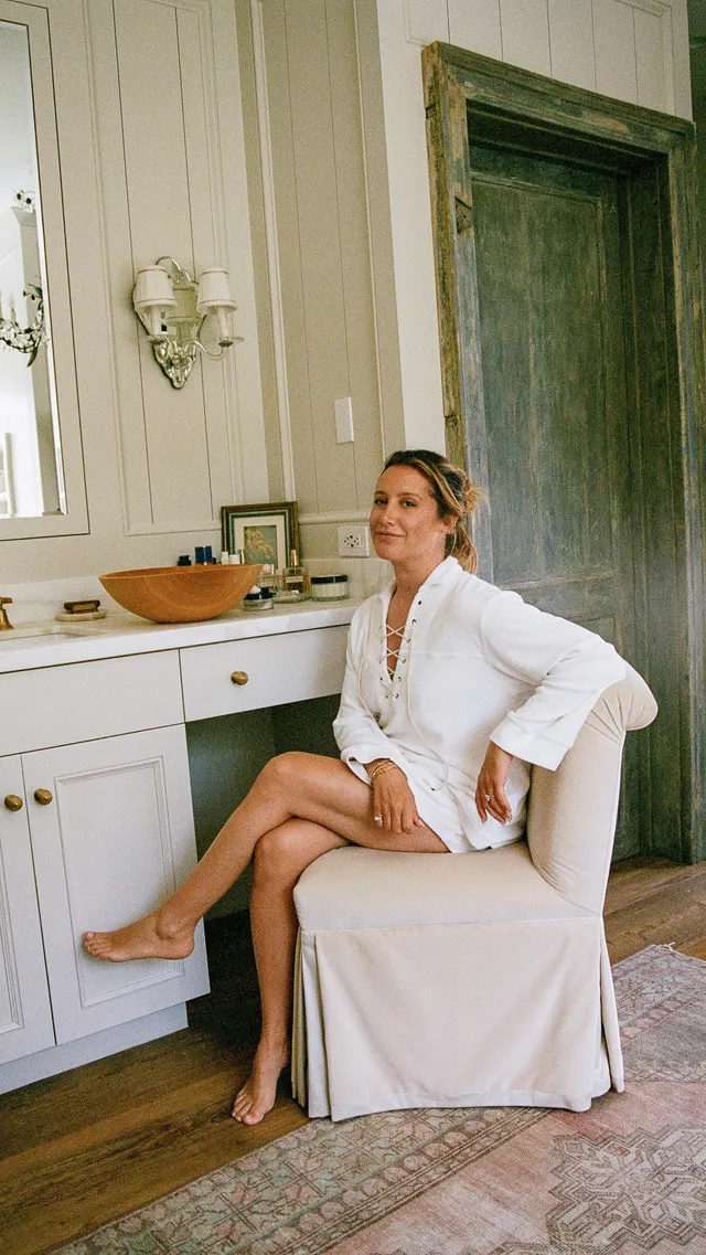 Ashley Tisdale sitting barefoot in a chair beside a dressing table.