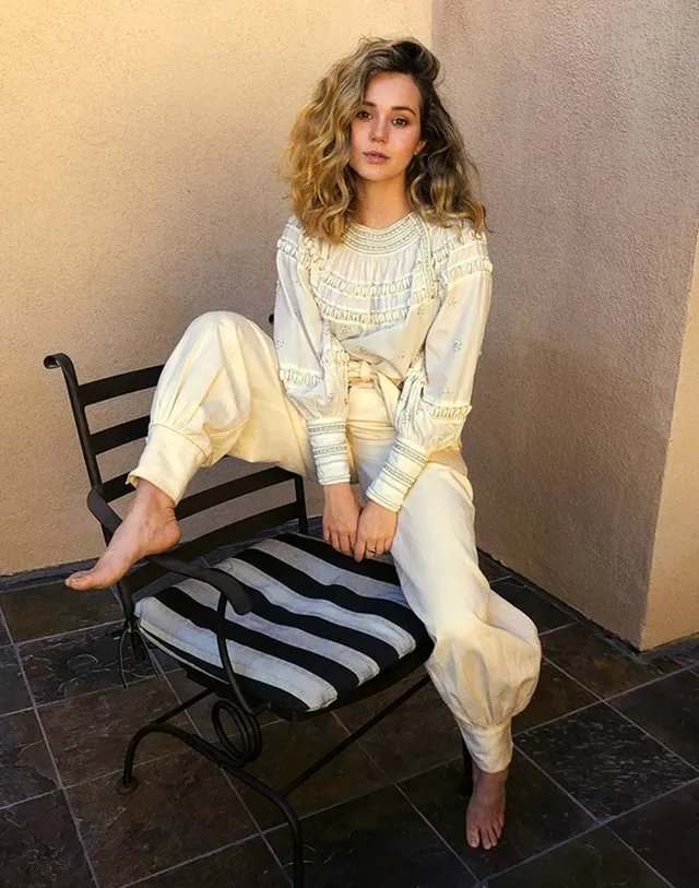 Brec Bassinger posing barefoot beside a chair in a pale striped outfit.