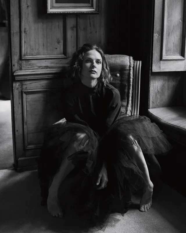 Rebecca Ferguson sitting in a dark dress in a monochrome portrait with bare feet visible.