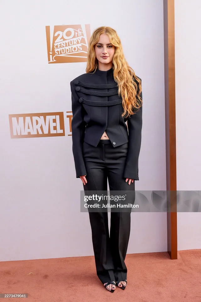 Grace Van Patten standing at an event in a black suit and heeled sandals.