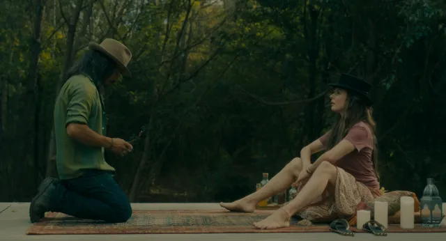 Rebecca Ferguson sitting barefoot on a wooden platform outdoors beside a companion.