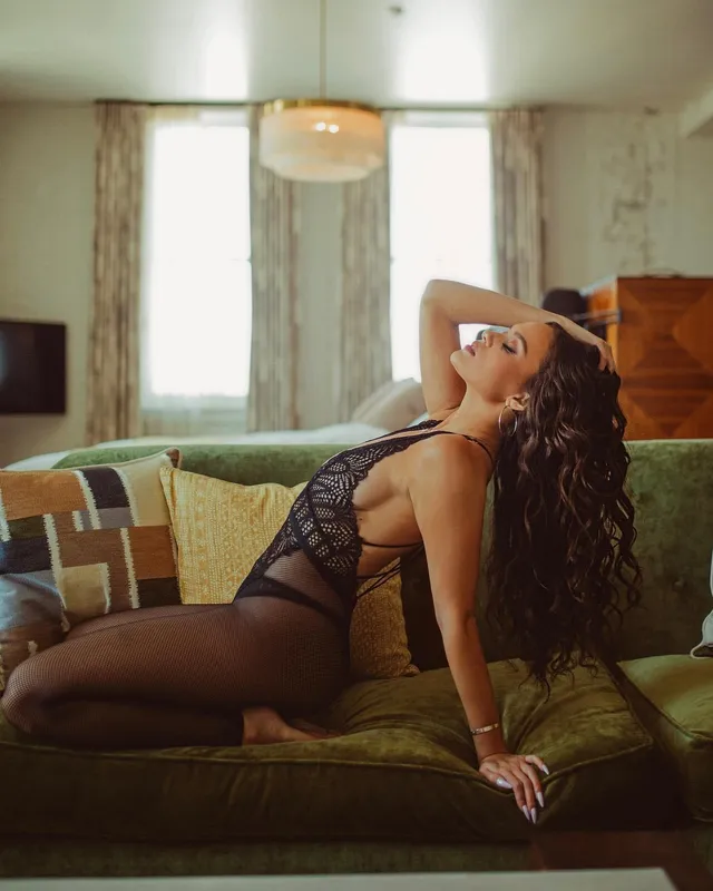 Madison Pettis reclining barefoot on a green sofa in a dark outfit.