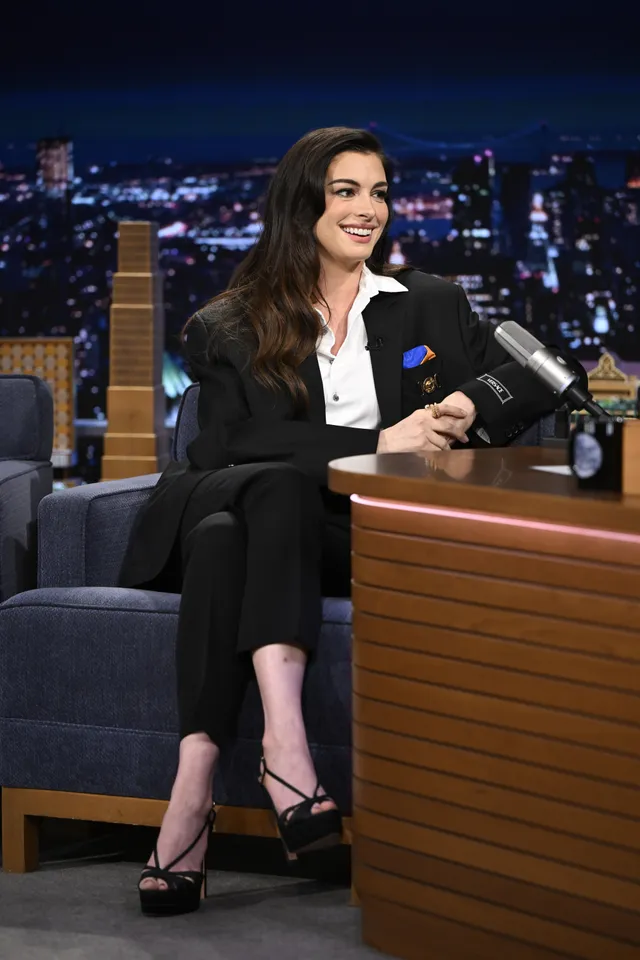 Anne Hathaway sitting during an interview in a dark suit and heels.