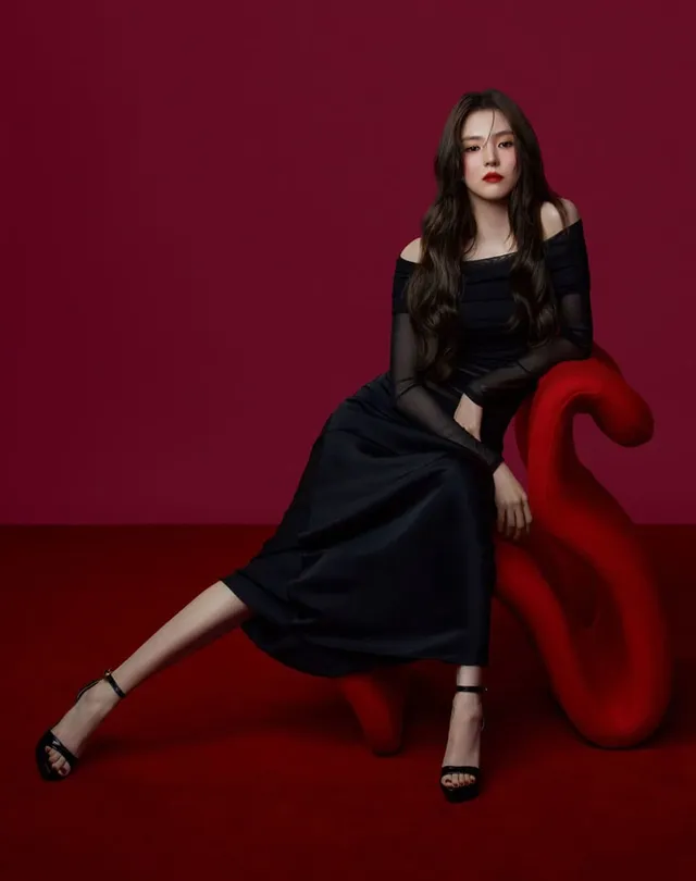 Han So-hee sitting in a black dress and heels against a red backdrop.