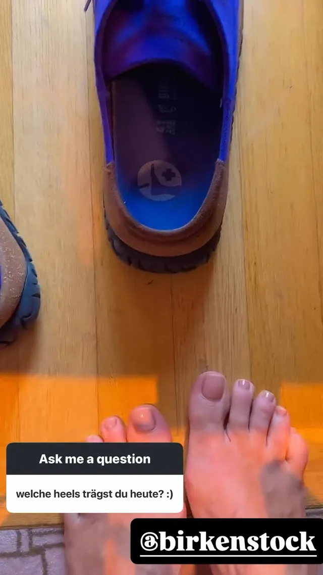 Heidi Klum resting her bare feet beside a blue flat shoe on a wooden floor.