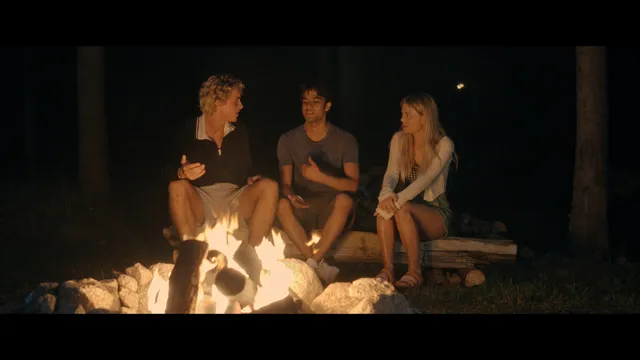 Emily Alyn Lind sitting barefoot with a group by an outdoor fire in a scene.