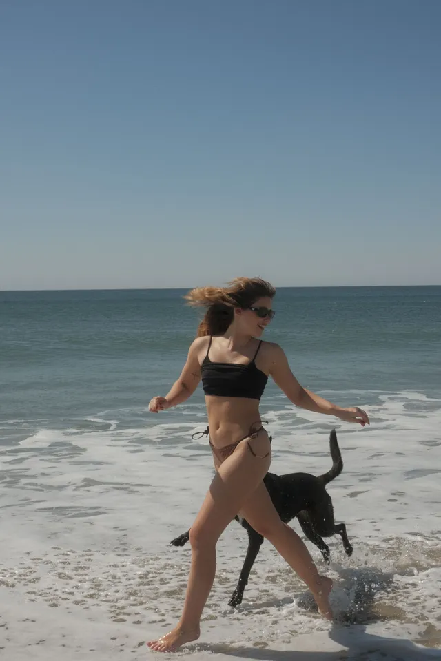 Nicole Wallace running barefoot beside a dog on a beach in a black bikini.