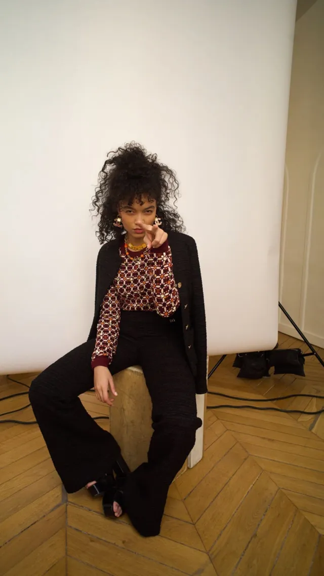 Whitney Peak sitting indoors in a patterned top, dark outfit and black heels.