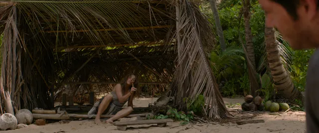 Rachel McAdams sitting barefoot beneath a palm shelter in a film scene.