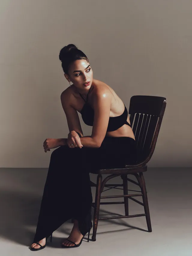 Melissa Barrera sitting on a wooden chair in a black outfit and heels.