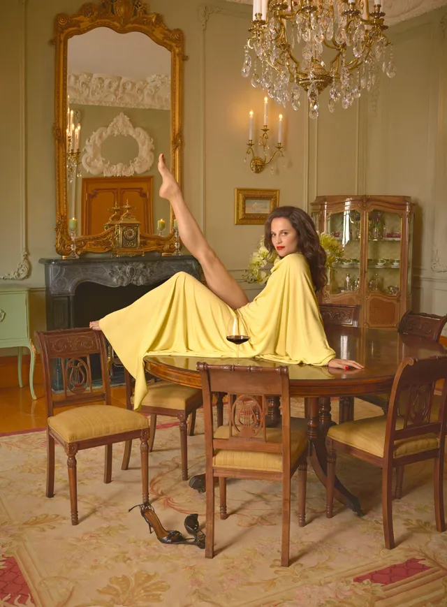 Alicia Vikander sitting on a dining table in a yellow outfit.