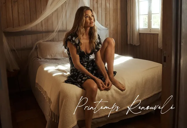Doutzen Kroes sitting barefoot on a bed in a dark floral dress.