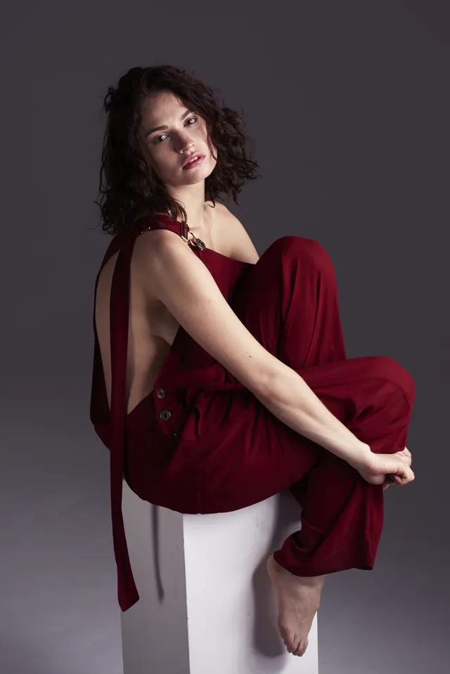 Lily James sitting barefoot on a white block in a burgundy outfit.