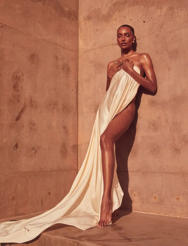 Jasmine Tookes standing barefoot in a flowing cream dress against a stone wall.