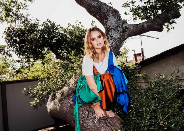 Debby Ryan sitting barefoot outdoors in a colorful outfit.