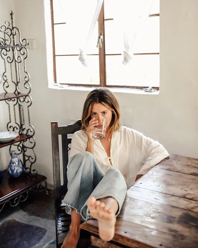 Ashley Tisdale sitting in a white top and jeans with her bare sole facing the camera.