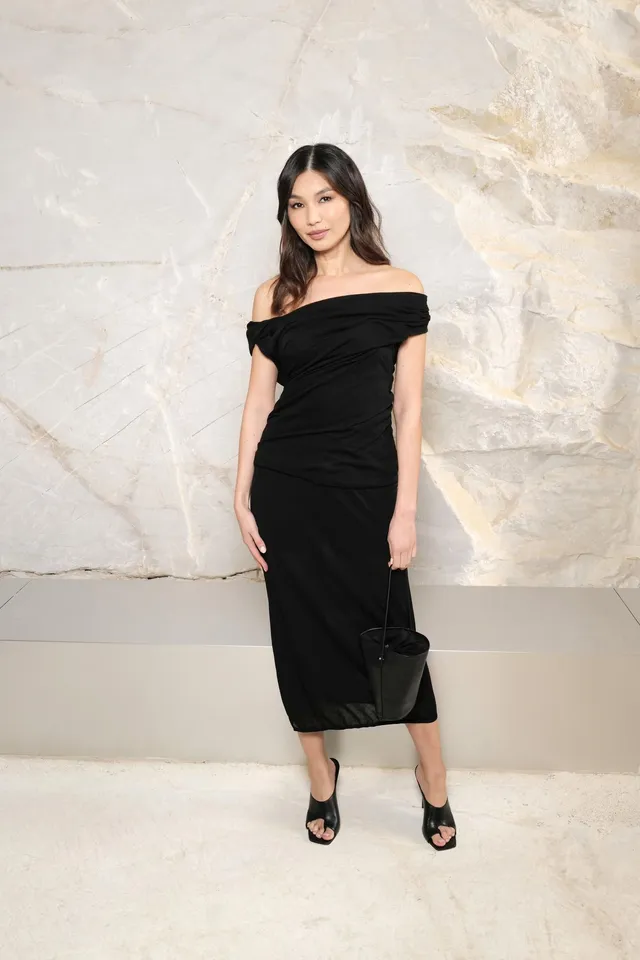 Gemma Chan posing against pale stone in a black off shoulder dress.
