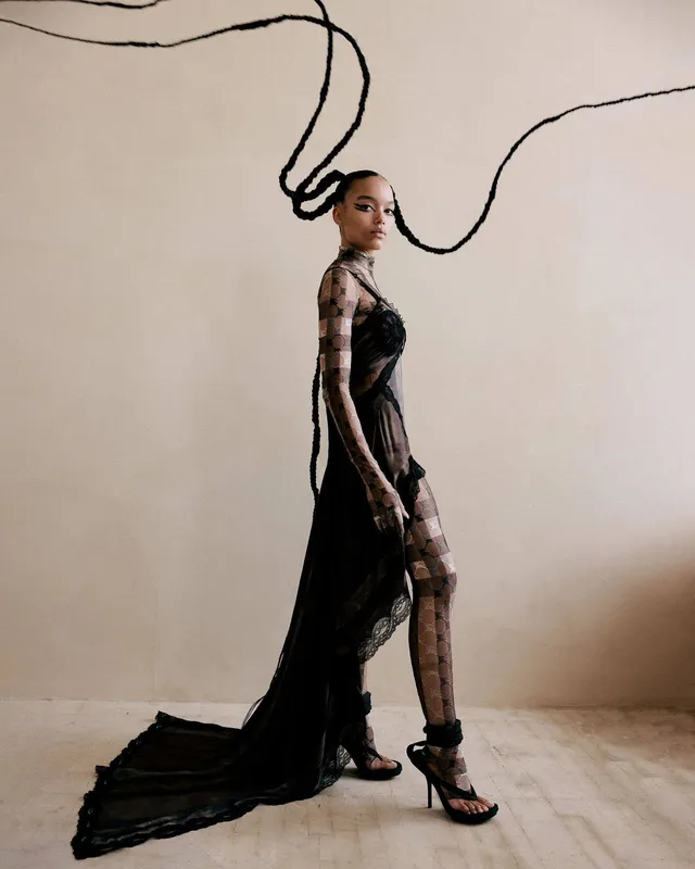 Whitney Peak posing in a long dark sheer dress and platform heels.