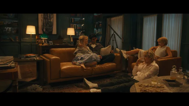 Emily Alyn Lind sitting on a sofa in a dress with bare feet visible in a scene.