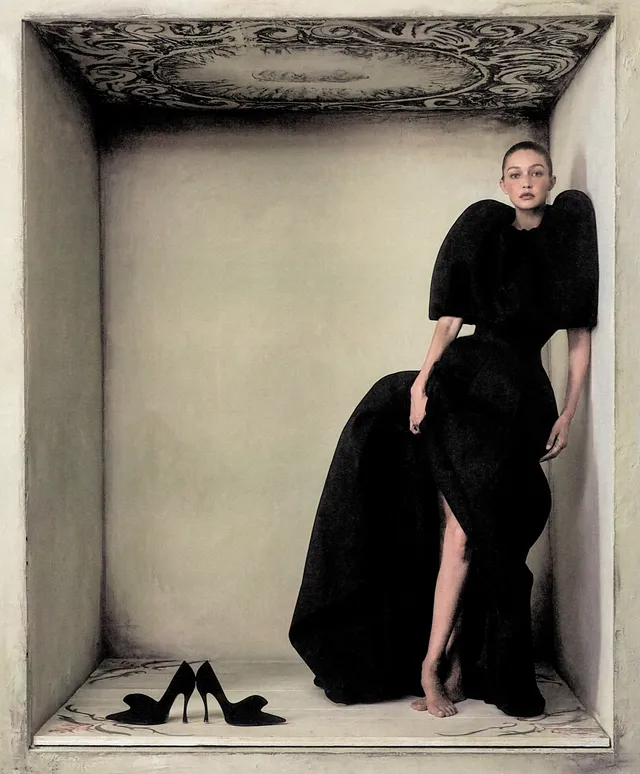 Gigi Hadid standing barefoot beside black shoes in a studio portrait.