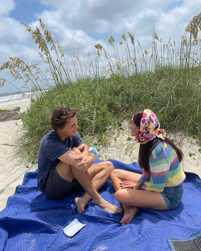 Lola Tung sitting barefoot on a beach blanket beside a man.
