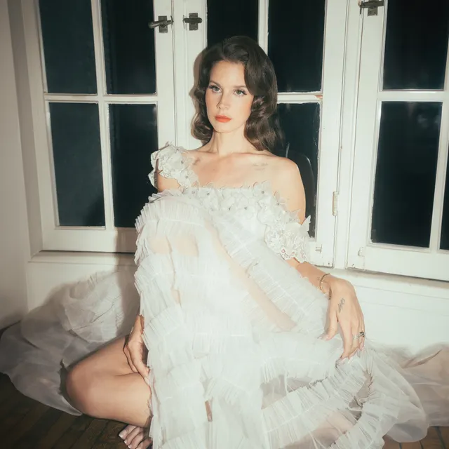 Lana Del Rey sitting barefoot in a pale dress beside a window.