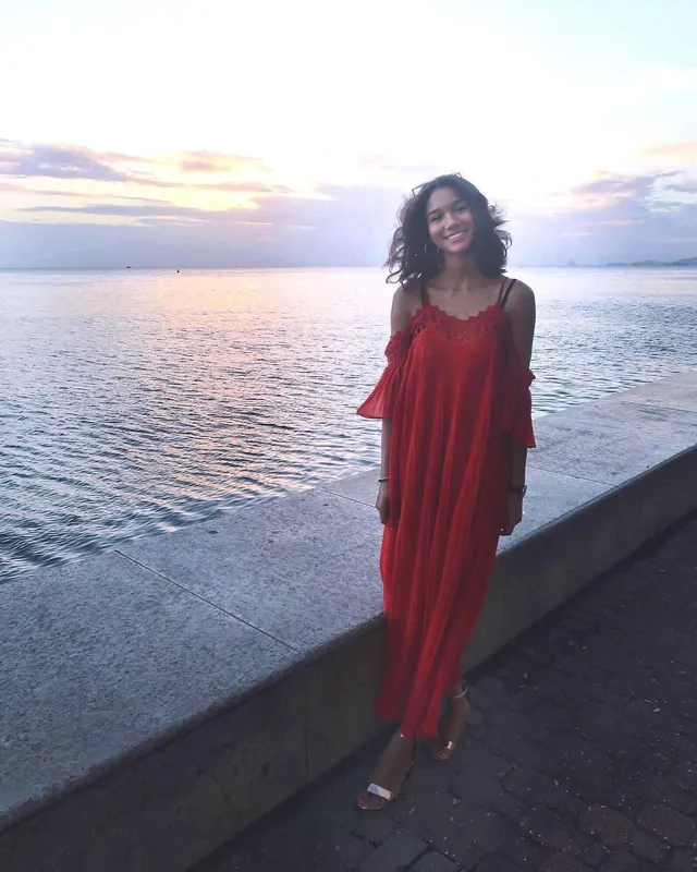 Greta Onieogou posing by the sea in a long red dress and flat sandals.