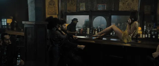 Rebecca Ferguson reclining barefoot across a bar counter in a film scene.