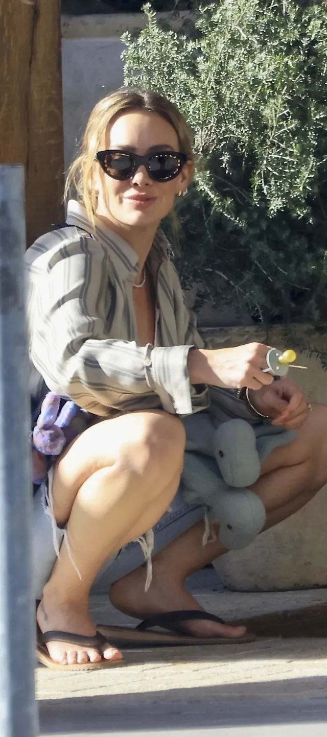 Hilary Duff crouching outdoors in sunglasses and flip-flops.