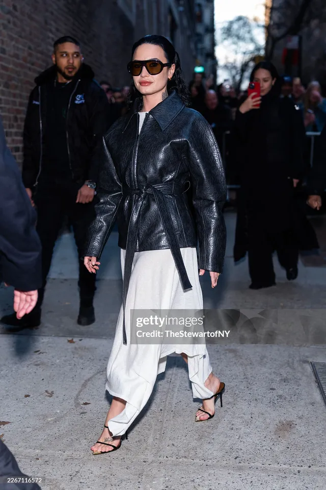 Demi Lovato walking outdoors in a leather jacket, white skirt and strappy heels.